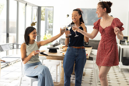 Laughing and toasting with wine glasses, three young women enjoy sunny afternoon indoors. Celebrating friendship and happiness, they create joyful memories in modern, stylish home settingの写真素材