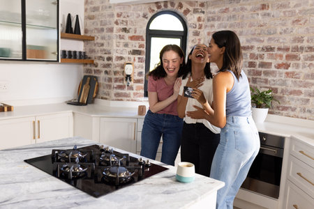 Laughing and embracing, three young women enjoy moment in modern kitchen. Friendship and joy fill air as they share stories and laughter over coffeeの写真素材