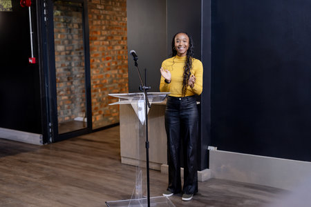 African American woman in mustard top clapping behind clear lectern with microphone in event space. Speaker, presentation, auditorium, modern, professional, inspiration, celebrationの写真素材