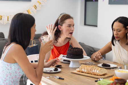 Celebrating with cake and drinks, three women gather around wooden table at home. Friends sharing...