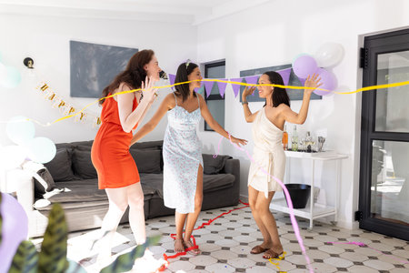 Dancing joyfully, three women in colorful dresses celebrate indoors with balloons and streamers. Friendship and happiness fill room as they enjoy lively party atmosphereの写真素材