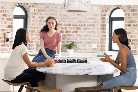 Three young women sitting around marble kitchen table, engaged in conversation. They are sharing stories and enjoying relaxed afternoon together in cozy, modern kitchenの写真素材
