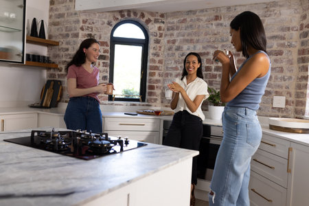 Laughing and chatting, three young women gather in cozy kitchen. They are enjoying each other's company, creating warm and joyful atmosphereの写真素材