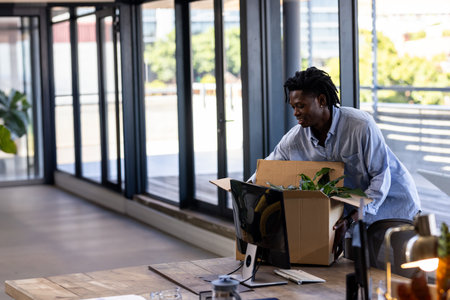 African American man unpacking potted plant from cardboard box in office by monitor, copy space. Professional, workspace, modern, interior, greenery, balcony, naturalの写真素材