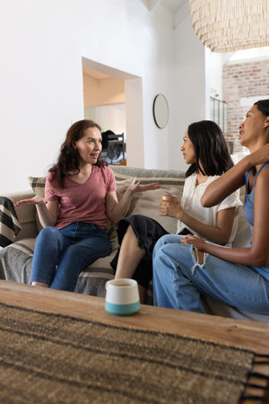 Chatting animatedly on cozy sofa, three young women enjoy relaxed afternoon. Sharing stories and laughter, they strengthen their friendship over warm drinks in stylish living roomの写真素材