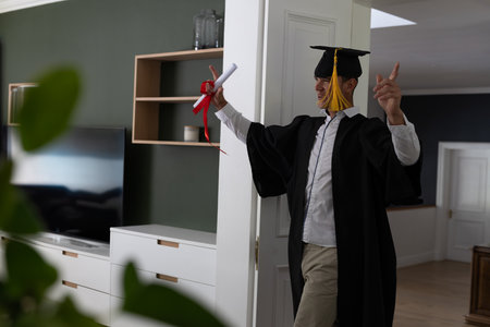 Male graduate raising arms holding diploma ribbon in home hallway with mortarboard cap, copy space. Celebration, achievement, success, modern, interior, accomplishment, prideの写真素材