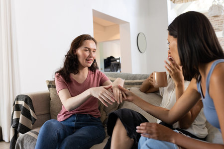 Laughing and chatting, three young women relax on cozy sofa at home. They share stories and enjoy each other's company, creating warm and friendly atmosphereの写真素材