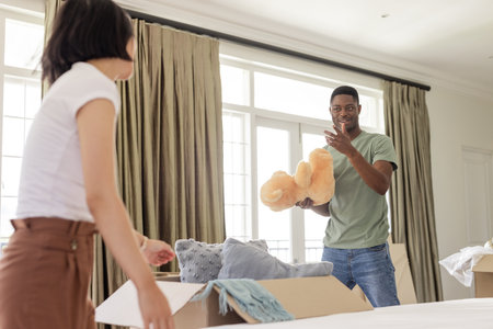 Couple unpacking boxes at home, smiling and holding teddy bear together. Moving, relocation, new...