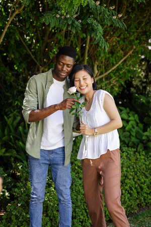 Couple enjoying romantic moment in garden, holding rose and smiling together. Romance, love, affection, togetherness, relationship, bondingの写真素材