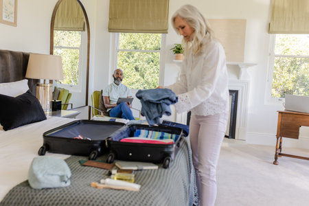 Woman packing suitcase in bedroom while man sitting on bed, preparing for trip. Travel, vacation, luggage, preparation, couple, getawayの写真素材