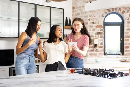 Three young women in casual outfits laughing and looking at photo in kitchen. They are sharing joyful moment, highlighting friendship and togetherness in cozy home settingの写真素材