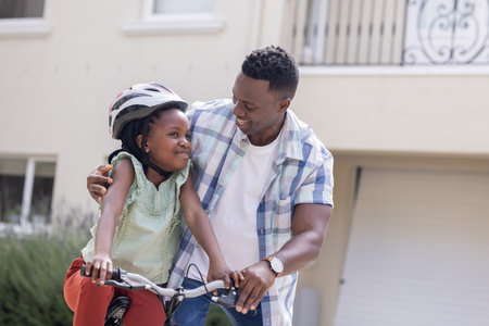 African American father teaching daughter to ride bicycle, both smiling outdoors. Family, parenting, bonding, cycling, fatherhood, childhoodの写真素材