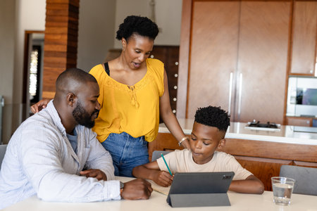 Family helping son with homework at kitchen table, using tablet and pencil. education, learning, teaching, digital device, parentingの写真素材