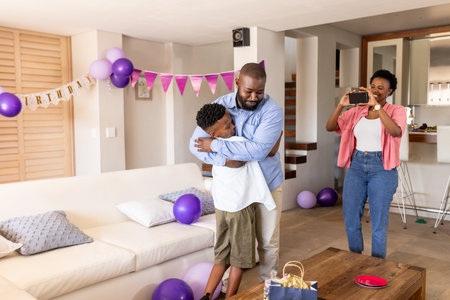 Celebrating birthday, father hugging son in living room while mother taking photo. Family,...
