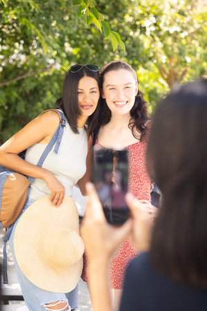 Two young women, one with long dark hair and other with light brown hair, pose happily outdoors on sunny day. They are enjoying moment together while friend captures their photo, highlighting friendship and joyの写真素材