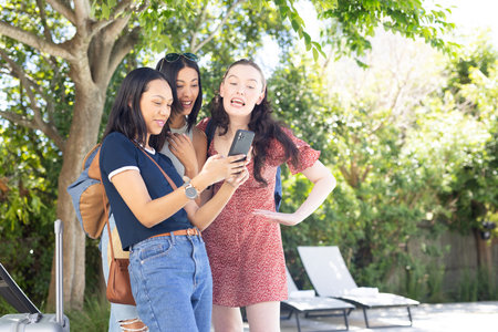 Three young women, one African American and two Caucasian, laughing while looking at smartphone outdoors. They are enjoying sunny day under tree, sharing joyful moments and friendshipの写真素材