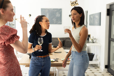 Laughing and chatting, three young women enjoy drinks at casual gathering. Celebrating friendship, they share stories and create joyful memories togetherの写真素材