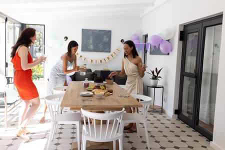 Setting up festive table, three women in colorful dresses prepare for celebration. Balloons and banner create joyful atmosphere in sunlit dining areaの写真素材