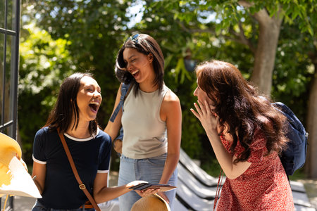 Laughing together, three young women enjoy sunny day outdoors with backpacks. Their joyful...