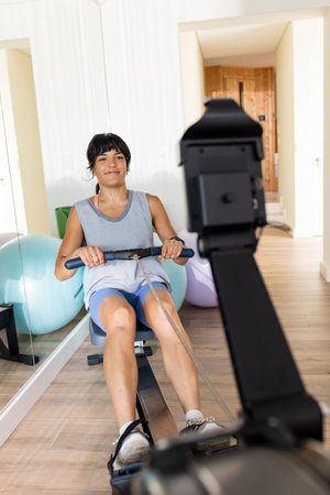 Woman exercising on rowing machine at home, smiling and staying fit. Fitness, exercise, workout,...