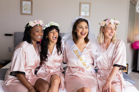 Bride-to-be with friends wearing robes and flower crowns, laughing on bed. Bachelorette, celebration, friendship, bridal party, floral, pajamasの写真素材