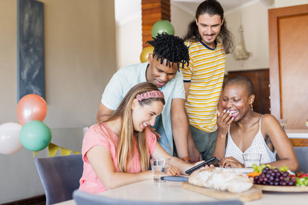 Diverse friends laughing and sharing moments around table with smartphone and snacks. Friendship,...
