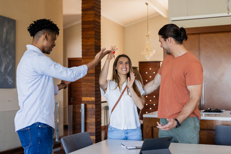 Friends celebrating success together at home, smiling and high-fiving around tablet. Celebration, friendship, happiness, high five, technology, indoorsの写真素材