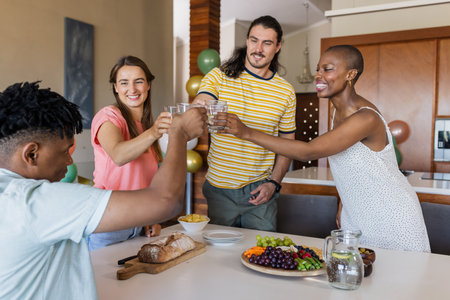 Friends toasting with water glasses around table with fruit platter in kitchen. Celebration,...
