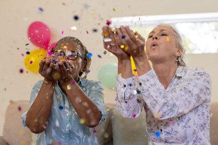 Blowing colorful confetti, senior women joyfully celebrating together at home, smiling. Celebration, festive, happiness, party, cheerful, elderの写真素材