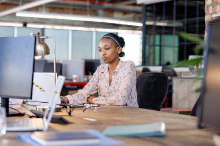 African American woman working on laptop in modern office, concentrating on task. business, professional, concentration, technology, workplace, corporateの写真素材