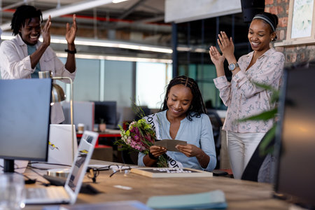 African American woman reading card with flowers, colleagues applauding in office. Celebration, appreciation, teamwork, diversity, corporate, coworkingの写真素材