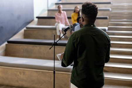 Presenting with microphone, African American man speaking to diverse audience in school hall. Public...