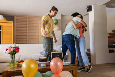 Friends celebrating at home, hugging and smiling with balloons and cake on table. Celebration, friendship, party, happiness, joy, festiveの写真素材