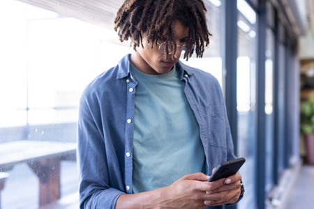 African American man using smartphone in modern office, concentrating on screen. Technology, concentration, professional, communicationの写真素材