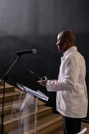 African American man presenting with microphone at podium in conference hall. Speaker, presentation, audience, public speaking, business, leadershipの写真素材