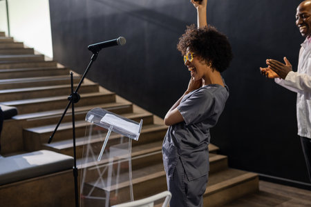 Woman celebrating success at podium while colleague applauding in conference hall. Victory,...