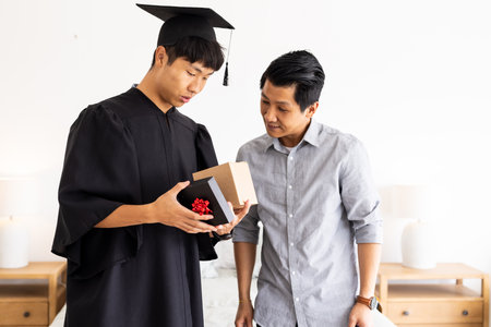 Graduate in cap and gown opening gift box with father at home, feeling surprised. Graduation, achievement, celebration, family, accomplishment, happinessの写真素材
