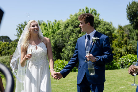 Bride and groom holding hands, smiling and celebrating outdoors with bubbles. Wedding, celebration, couple, romance, love, festivityの写真素材