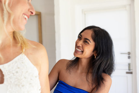 Smiling woman in blue dress helping bride with wedding gown at home. preparation, friendship, celebration, dressing, assistance, bridalの写真素材
