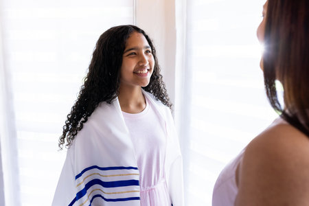Young girl wearing prayer shawl smiling at woman during bar mitzvah. tradition, spirituality,...
