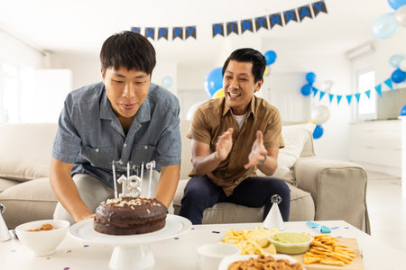 Blowing candles on birthday cake, Asian man with friend clapping joyfully. Celebration, party, friends, happiness, tradition, eventの写真素材