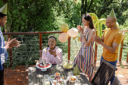 Friends clapping as African American woman blows candles celebrating birthday outdoors. Celebration, friendship, happiness, diversity, birthday party, cakeの写真素材