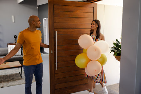 Welcoming African American woman with balloons entering home, greeted by smiling man. Celebration,...