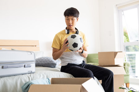Unpacking boxes in bedroom, young man holding soccer ball with headphones. moving, casual, lifestyle, home, maleの写真素材