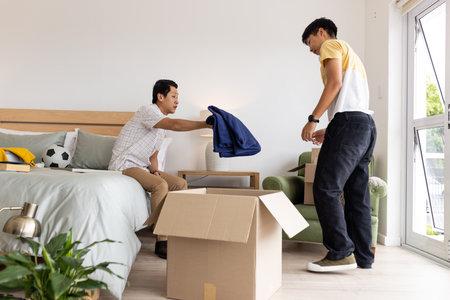 Father and son unpacking boxes in bedroom, organizing clothes together. Family, bonding, teamwork, parenting, domestic, lifestyleの写真素材