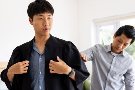 Young man adjusting graduation gown while friend helps at home, feeling proud. achievement, education, celebration, friendship, academic, milestoneの写真素材