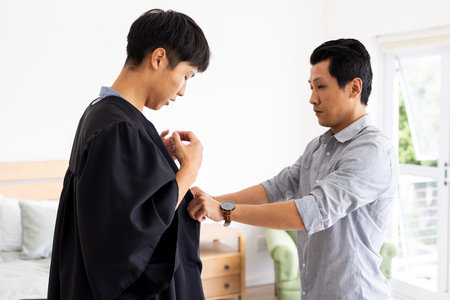 Father helping son adjust graduation gown at home, preparing for ceremony. family, celebration, achievement, support, fatherhood, educationの写真素材