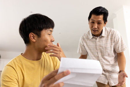 Father discussing important document with son at home, both appearing thoughtful. Parenting, communication, family, bonding, fatherhood, relationshipの写真素材