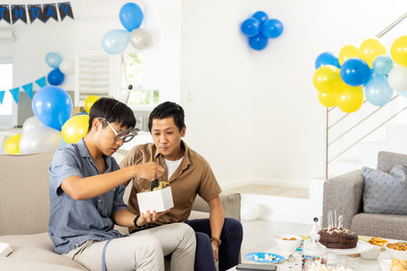 Father and son celebrating birthday at home, opening gift with balloons around. Celebration, family, happiness, childhood, surprise, family timeの写真素材