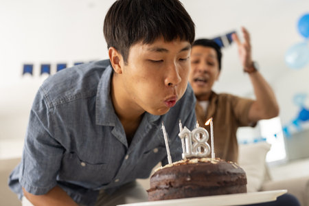 Young man blowing out candles on birthday cake while dad celebrates joyfully. Celebration, family, happiness, party, father, sonの写真素材